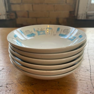 May include: A stack of six white ceramic bowls with a light blue mid-century modern design. The bowls are stacked on a wooden surface, with a window and brick wall in the background.