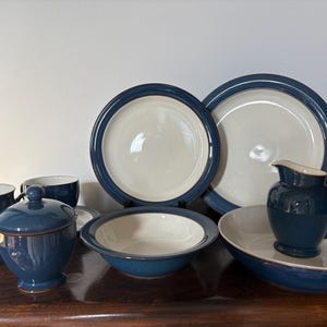 May include: A ceramic dinnerware set featuring a two-tone design with a deep navy blue rim and a cream-colored center. The collection includes plates, a bowl, a lidded sugar bowl, a creamer pitcher, and mugs, all with a smooth, glossy finish.
