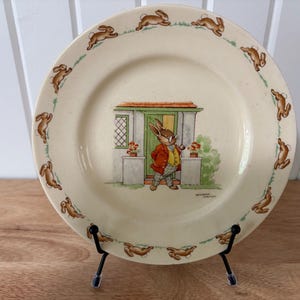Op de afbeelding: Een vintage keramische bord met een grillige illustratie van een konijn in een rode jas en groene broek voor een huis. Het bord heeft een crèmekleurige achtergrond met een rand van rennende konijnen. Het bord wordt gepresenteerd op een zwarte metalen standaard.