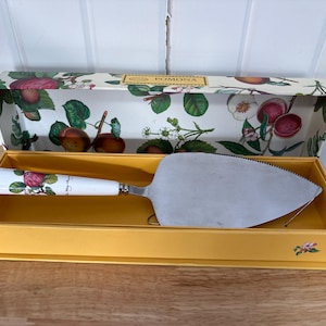 May include: A cake server with a stainless steel blade and a white handle decorated with red apples and green leaves. The server is in a yellow box with a floral design and the word "POMONA".