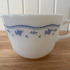 May include: White teacup with a blue floral design. The cup has a curved shape and a handle. A blue scalloped border with floral and leaf patterns decorates the top of the cup. The teacup is likely vintage or retro.