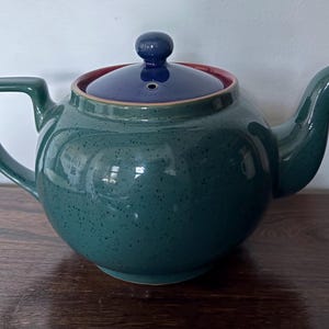 Könnte beinhalten: Eine Teekanne aus Keramik in Türkis mit gesprenkelter Oberfläche. Der Deckel ist marineblau mit einem kleinen Knauf, der Rand ist rot. Sie hat einen gebogenen Ausguss und einen eckigen Griff. Ein klassisches Tee-Objekt.