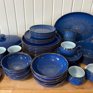May include: A full set of blue ceramic tableware featuring a floral pattern. The collection includes plates, bowls, cups, saucers, a serving platter, and a lidded casserole dish. The edges have a brown trim.