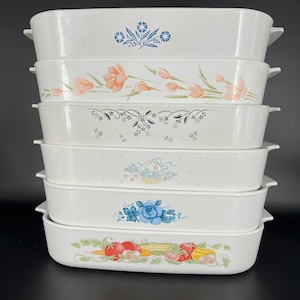 May include: A stack of six white ceramic baking dishes with different floral and patterned designs. The dishes are stacked in a pyramid shape, with the largest dish at the bottom and the smallest dish at the top.