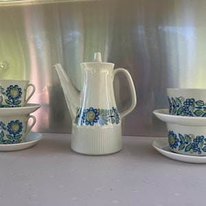 May include: A white teapot and four white teacups with blue and green floral patterns. The teapot has a spout and a handle. The teacups have handles and are stacked in pairs.