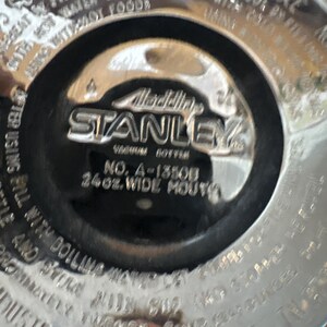May include: Close-up of the bottom of a Stanley vacuum bottle. The center features the Stanley logo and text that reads "Vacuum Bottle" and "24oz. Wide Mouth". The bottle is silver and black.