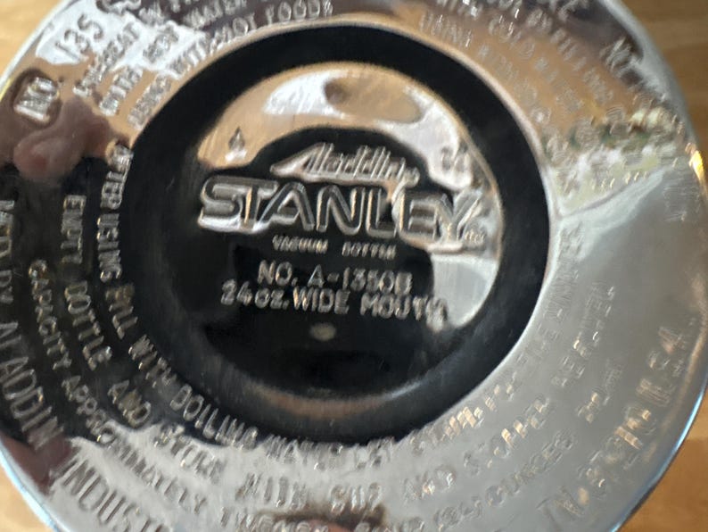 May include: Close-up of the bottom of a Stanley vacuum bottle. The center features the Stanley logo and text that reads "Vacuum Bottle" and "24oz. Wide Mouth". The bottle is silver and black.