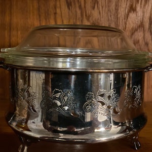 May include: A silver-plated serving dish with a clear glass lid. The dish features ornate floral detailing and small feet. The handles are curved. The lid has a rounded shape. The dish is likely used for serving food.