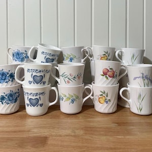 Pode incluir: Uma coleção de canecas de cerâmica branca com vários desenhos florais e de frutas. As canecas apresentam rosas azuis, corações e outros padrões botânicos. Algumas canecas têm um design texturizado e canelado.