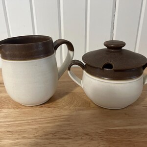 May include: A brown and white ceramic creamer and sugar bowl set. The creamer has a brown rim and a handle. The sugar bowl has a brown lid with a small spout.