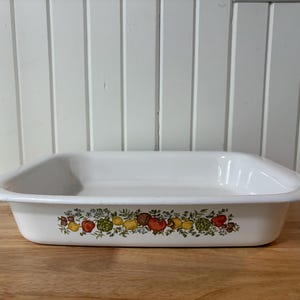 May include: A rectangular white enamel baking dish with a wide rim. The exterior features a colourful horizontal band of illustrations depicting various vegetables including red tomatoes, yellow peppers, green artichokes, and brown mushrooms.