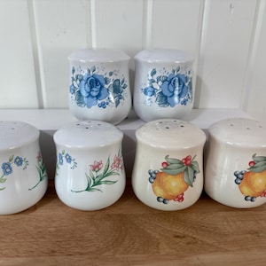 May include: A collection of six ceramic salt and pepper shakers. Two shakers feature blue rose designs, two have floral patterns, and two display yellow fruit illustrations. The shakers have white lids with multiple holes.
