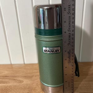 May include: A vintage green Aladdin Stanley thermos with a silver lid and handle. The thermos has a green body with the "Aladdin Stanley" logo. The thermos is approximately 10 inches tall.