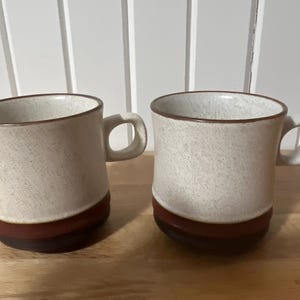 May include: Two white ceramic mugs with brown rims and brown bands around the bottom. The mugs have a speckled texture.