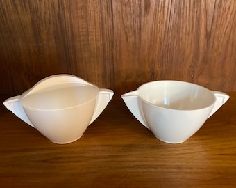 May include: A white ceramic creamer and sugar bowl set with a modern design. The creamer has a spout and the sugar bowl has a lid.