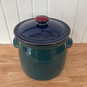 Puede incluir: Una olla de cerámica con tapa, con cuerpo verde oscuro y tapa azul marino. La tapa tiene un pequeño asa redonda de color burdeos. La olla tiene dos asas laterales y está sobre una superficie de madera. La olla mide unos 18 cm de altura.