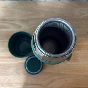 May include: An overhead view of a green and silver insulated thermos with a matching cup and lid. The thermos is open, revealing the interior. The items are on a light brown wooden surface.