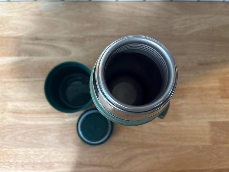 May include: An overhead view of a green and silver insulated thermos with a matching cup and lid. The thermos is open, revealing the interior. The items are on a light brown wooden surface.