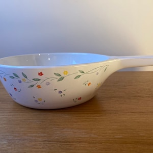 May include: White ceramic serving bowl with a floral pattern of small, colorful flowers and green leaves. The bowl has a handle.