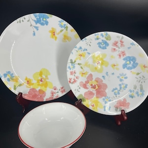 May include: A white ceramic dinner set with a floral pattern in yellow, blue, and pink. The set includes a large plate, a smaller plate, and a bowl with a red rim. The plates and bowl are displayed on a dark surface.