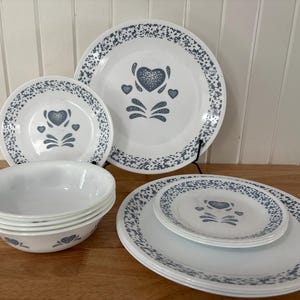May include: A white dinner service with a heart motif in the centre of each item. The plates and bowls have a speckled blue border. The set includes dinner plates, side plates, and bowls, ideal for serving meals.