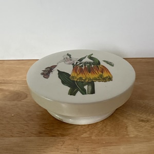 May include: A cream-coloured, round trinket box with a floral design. The lid features a detailed illustration of a yellow and orange flower with green leaves and a butterfly. The box sits on a short, rounded base.