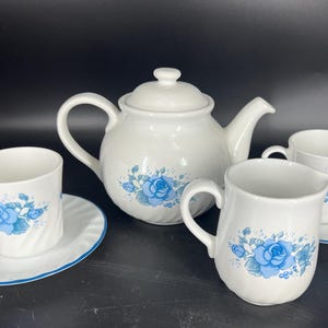 May include: A white teapot and three cups with blue floral designs. The teapot has a lid and a spout. The cups have handles. The cups and teapot are on a white background.