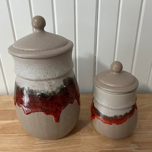 May include: Two ceramic canisters with lids. The canisters are brown with a white speckled glaze and a red and brown glaze drip design. The lids are brown with a white speckled glaze.