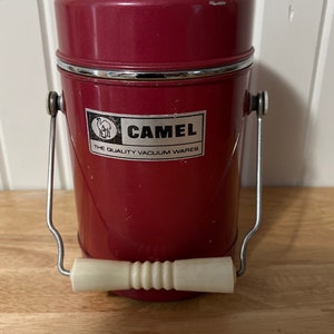 Puede incluir: Un recipiente de vacío cilíndrico de color rojo con un asa metálica giratoria y un agarre de plástico blanco. El frente presenta una etiqueta plateada con un gráfico de camello, el texto CAMEL y THE QUALITY VACUUM WARES. Tapa de metal rojo.