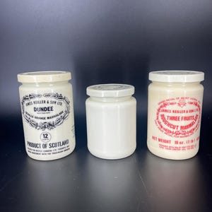 May include: Three vintage marmalade jars with white lids. The jars have labels with text, including "Dundee Seville Orange Marmalade" and "Three Fruits Roughcut Marmalade." The jars are cream-colored and sit on a dark surface.
