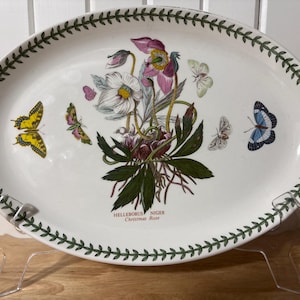 May include: Oval serving platter with a botanical design. The white ceramic platter features a central floral arrangement with pink and white flowers, green leaves, and butterflies. The rim is decorated with a green leaf pattern. Text on the platter reads "Helleborus Niger Christmas Rose."