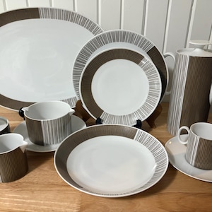 Op de afbeelding: Een compleet wit en bruin gestreept porseleinen servies. De set bevat een grote ovale schaal, borden, kopjes, schotels, een melkkan, een suikerpot en een koffiepot. De bruine strepen zijn verticaal op de koffiepot en de kopjes.
