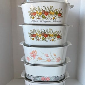 Pode incluir: Uma pilha de travessas brancas com tampas de vidro. Os dois pratos superiores apresentam um design colorido de vegetais e ervas com texto. Outros pratos têm padrões florais em rosa, laranja e azul.