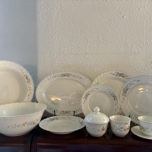 Könnte beinhalten: Eine Sammlung cremefarbener Porzellan-Geschirrteile mit floralen und blauen Verzierungen. Das Set umfasst Teller, Schalen, eine Servierplatte, eine Zuckerdose und Teetassen. Das Blumenmuster zeigt rosa, gelbe und blaue Blumen.