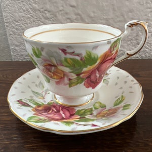 May include: A white teacup and saucer with a floral pattern featuring pink, yellow, and green roses. The cup and saucer have a gold trim.