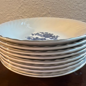May include: A stack of ten white ceramic bowls with a blue floral design and a silver rim.