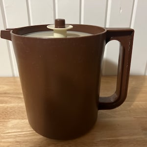 Può includere: Una brocca di plastica marrone con manico e coperchio. La brocca ha un beccuccio per versare e un coperchio color crema con un pomello marrone. La brocca è su una superficie di legno.