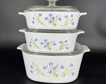 May include: Stack of three white casserole dishes with glass lids. Each dish features a blue floral design with green stems and leaves. The dishes are of varying sizes, with clear glass lids and handles.