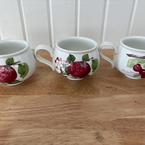 Könnte beinhalten: Drei weiße Keramikbecher mit gebogenen Griffen. Jeder Becher zeigt ein anderes Fruchtdesign: einen roten Apfel, Apfelblüten und Kirschen. Grüne Blätter und Stiele begleiten die Fruchtdarstellungen. Die Becher stehen auf einer Holzoberfläche.