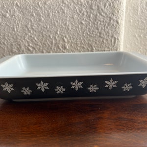 May include: A black baking dish with a white snowflake design. The dish is made of ceramic and has a white interior.