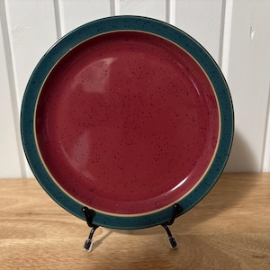 May include: A round ceramic plate with a burgundy centre speckled with dark spots. The plate has a teal blue rim and a beige border. The plate is displayed on a black metal stand, suitable for serving food.
