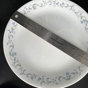Może przedstawiać: Biały ceramiczny talerz obiadowy z zielonym i niebieskim wzorem kwiatowym wok&oacute;ł krawędzi. Talerz ma średnicę 23 cm.