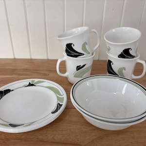 Peut inclure: Un service de table en céramique blanche avec un motif floral noir et vert. L'ensemble comprend des tasses, des bols et des assiettes ovales. Les tasses ont des anses et les bols ont un bord noir et vert.