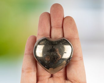 May include: A polished, metallic pyrite heart-shaped stone. The heart is held in an open hand, reflecting light with a golden sheen. The background is a soft gradient of green and white.