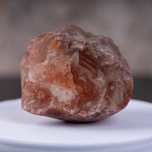 May include: A rough, reddish-brown agate stone with a translucent, layered interior. The stone is sitting on a white surface.