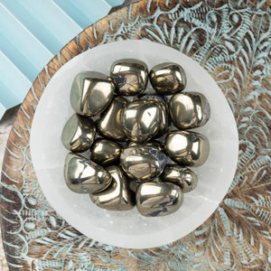 May include: A white bowl filled with a collection of smooth, golden pyrite tumbled stones. The bowl is sitting on a wooden surface with intricate carvings.
