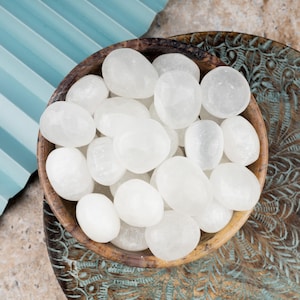 May include: A bowl filled with white selenite tumbled stones. The bowl is made of wood and has a natural finish. The stones are smooth and polished.