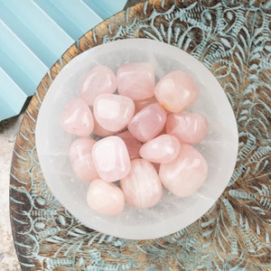 May include: A white bowl filled with pink rose quartz tumbled stones. The bowl is sitting on a wooden surface with a blue and white patterned design.