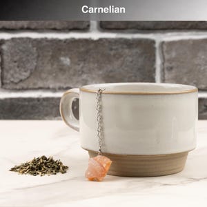 May include: A white ceramic mug with a brown base and rim, paired with a carnelian tea infuser. The infuser features a chain and a raw, orange-colored carnelian stone. A small pile of loose green tea leaves sits beside the mug. The word "Carnelian" is at the top.