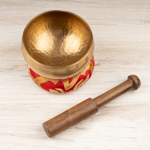 May include: A gold-coloured metal singing bowl with a hammered texture, resting on a circular red fabric cushion featuring gold embroidered patterns. A cylindrical wooden mallet with a smooth finish is positioned next to the bowl.
